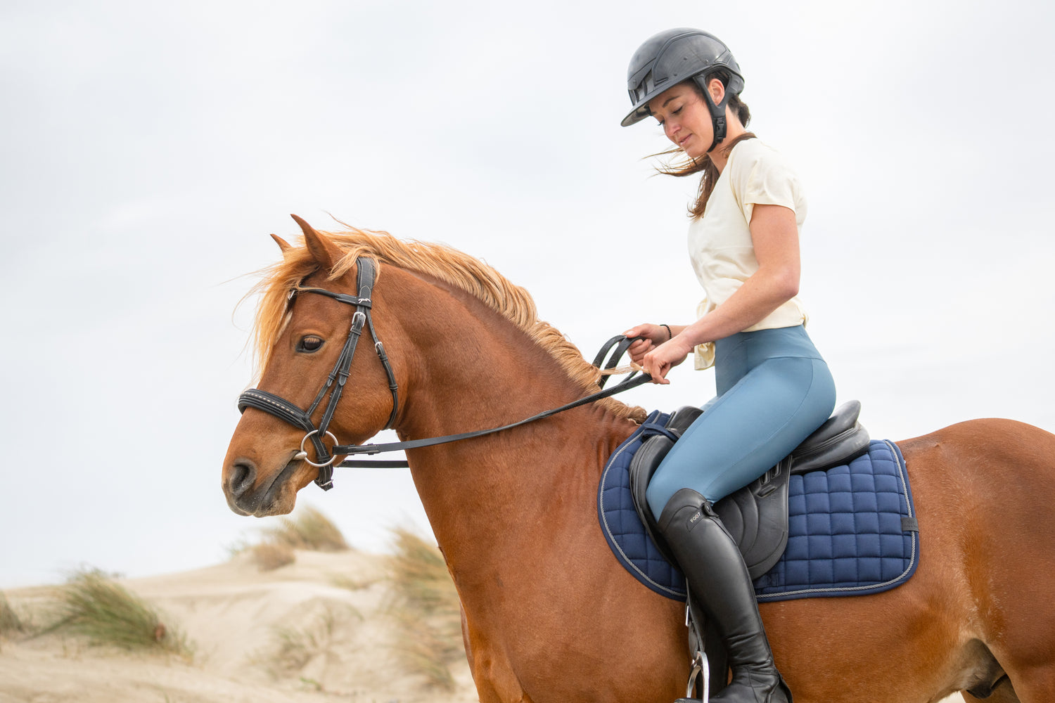 A woman riding a chestnut horse on a sandy beach, wearing blue leggings and a helmet