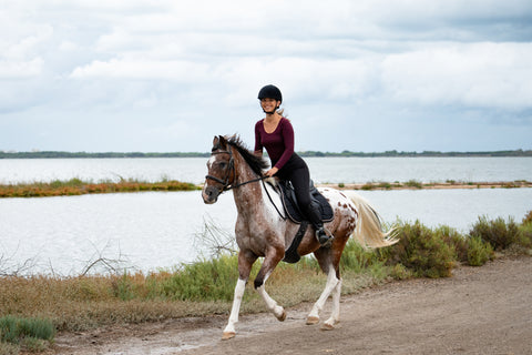 A woman riding a spotted horse along a path by a lake, wearing a helmet and dark riding attire