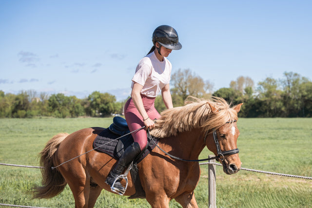 A person wearing a helmet and riding a brown horse in an open grassy field on a sunny day