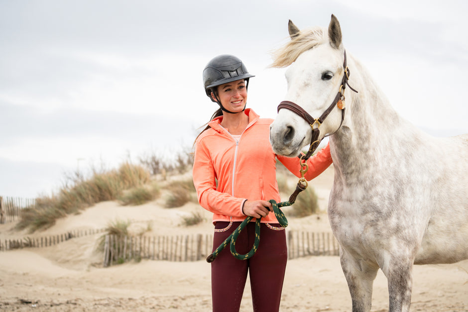 A woman in an orange jacket and riding helmet smiling while holding a white horse's lead rope on a sandy beach