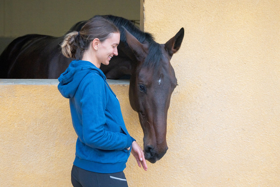 Person in a blue hoodie standing beside a horse peeking through a window