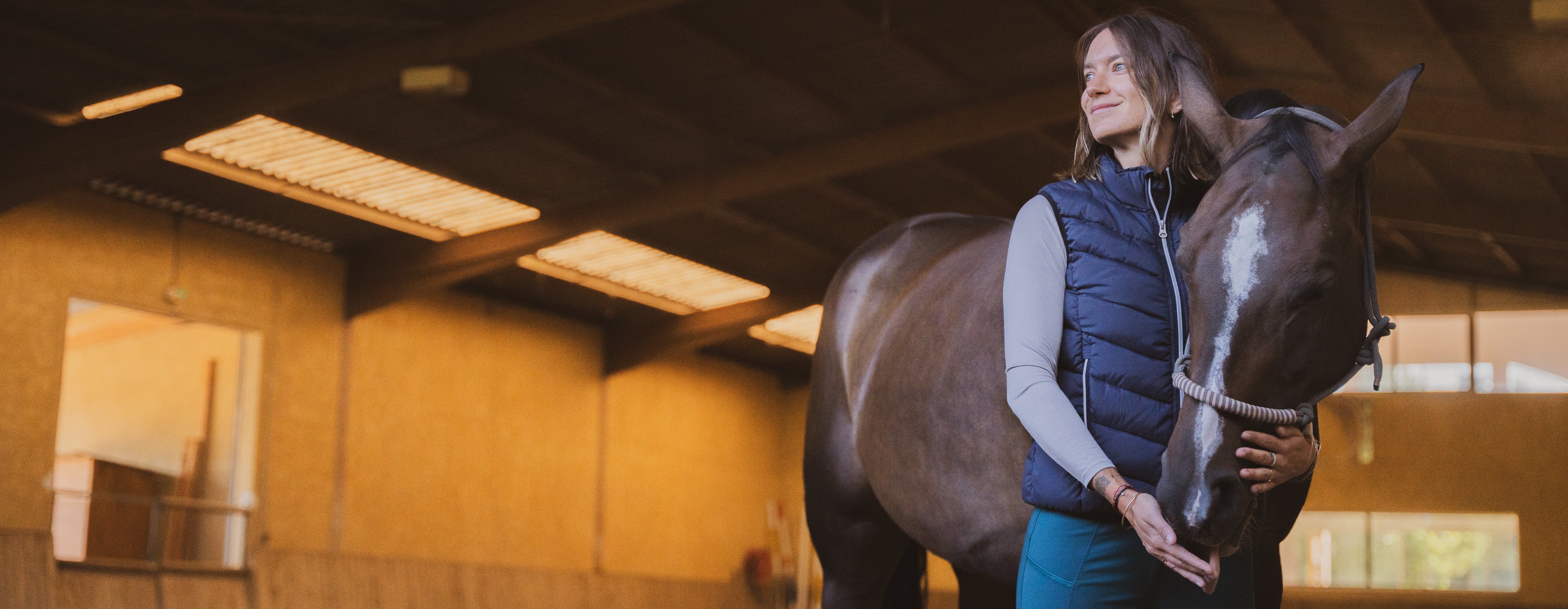 A woman in an indoor stable, smiling and standing close to a dark horse