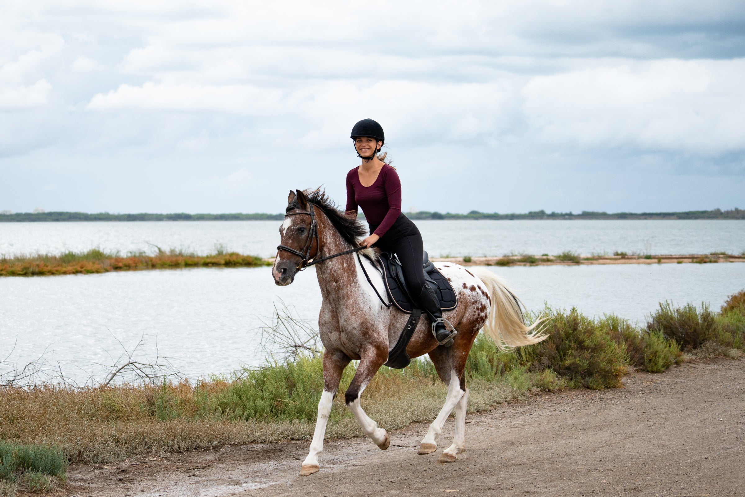 A person riding a spotted horse along a dirt path by a body of water, wearing a helmet and a long-sleeved top